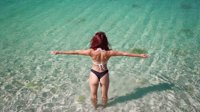 Back View, Beautiful Slim Woman with Red Hair in Swimsuit Raises His Hands Up from Happiness and Freedom Standing in the Transparent Turquoise Sea. Summer Vacation and Travel. Slow motion. Top view