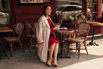 Attractive brunette woman in red dress, beige trench coat and beret smiles, sits in street cafe and enjoys coffee.