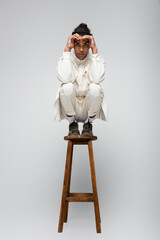 stylish african american man in white suit and sneakers posing on high stool on grey