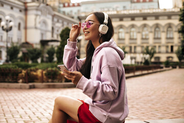 Fototapeta na wymiar Happy young brunette Asian girl in stylish hoodie, pink sunglasses and headphones listens to music and holds phone outside.