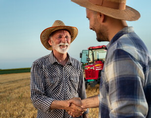 Male farmers standing in field in front of tractor shaking hands.