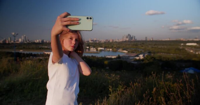 Blonde teen taking selfie in park. Closeup of teenager searching best pose for selfie portrait. Trendy girl taking selfie photo on smart phone