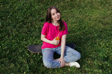 Smiling Young woman in pink t-shirt sitting on skateboard on green grass and holding bottle of lemonade. Summer activity, healthy lifestyle, sport, fitness. Female skater. Having fun outdoors