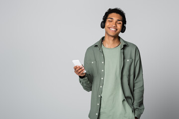 african american guy with smartphone listening music while holding hand in pocket isolated on grey