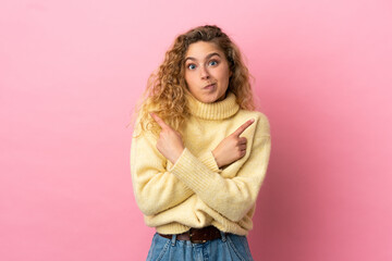 Young blonde woman isolated on pink background pointing to the laterals having doubts