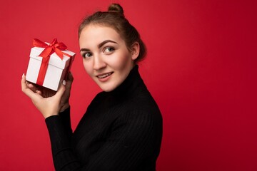 Portrait photo shot of beautiful happy thoughtful young brunette woman isolated over red background wall wearing black sweater holding white gift box with red ribbon and looking to the side