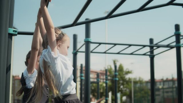 Two little schoolgirls jumping onto the monkey bars at a playground in schoolyard. Schoolmates wearing school uniform and play during school break.