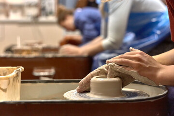 Hands of a ceramist while she is working on a potter's wheel against the background of other people working with clay
