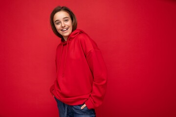 Fototapeta premium Shot of beautiful happy cool smiling brunette little girl wearing trendy red hoodie standing isolated over red background wall looking at camera