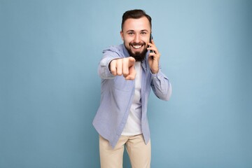 Photo shot of positive smiling good looking young brunet bearded man wearing casual blue shirt and white t-shirt poising isolated on blue background with empty space holding in hand and communicating