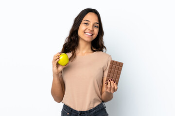 Young woman over isolated white background taking a chocolate tablet in one hand and an apple in...