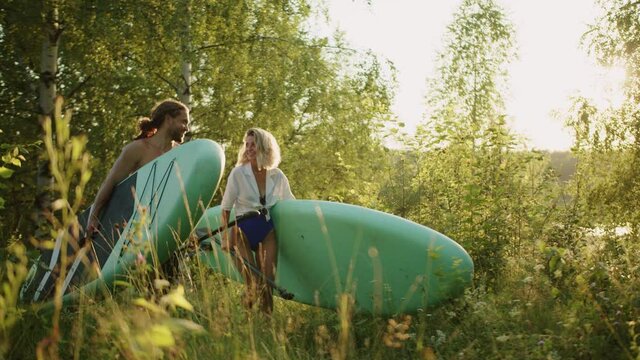 Surfers happy couple carrying surfing boards with paddles. Sports family smiling, walking with paddleboards. Professional trainer and beginner with surfboards going to river. Summer sport, nature.