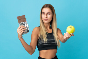 Young blonde woman isolated on blue background having doubts while taking a chocolate tablet in one...
