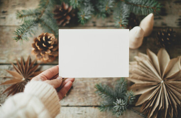 Christmas card mock up. Hand holding empty greeting card on background of christmas paper stars, wooden tree, pine branches and cones on rustic wood.  Space for text. Seasons greetings template - Powered by Adobe