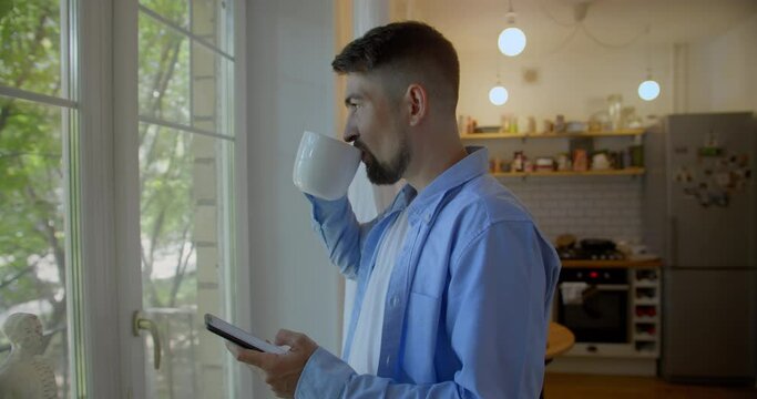 Young man talking on a mobile phone in the morning at a window with coffee
