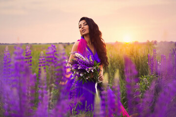 A brunette girl in a gradient haute couture dress in blue, pink and yellow colors standing among a blooming lupine field. A magical romantic sunset portrait.