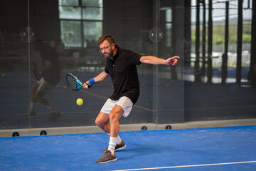 Man playing padel in a blue grass padel court indoor - Young sporty boy padel player hitting ball...