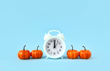 Alarm clock surrounded by four toy orange pumpkins on light blue background. Halloween time...