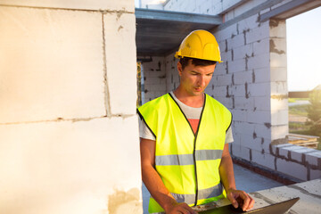 Construction engineer on the construction site of a house made of porous concrete blocks works at a computer in a reflective safety vest and hardhat. Design, construction, drawings project, check