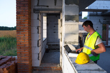 Fototapeta na wymiar Construction engineer on the construction site of a house made of porous concrete blocks works at a computer in a reflective safety vest and hardhat. Design, construction, drawings project, check