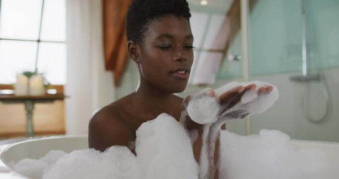 Smiling african american attractive woman taking bath and blowing out foam in bathroom