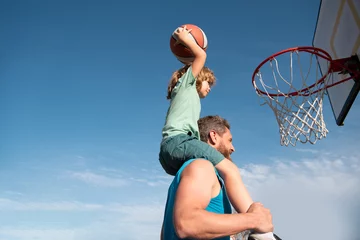  Father and son playing basketball. Sports Concept. Boy child sitting on the dad shoulders, throwing basketball ball into basket, side view on sky background with copy space. © Volodymyr
