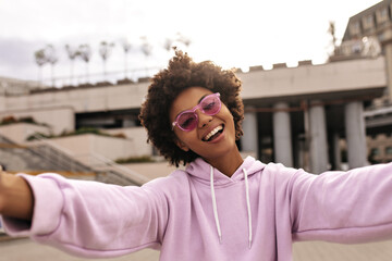 Attractive charming woman in pink sunglasses and purple hoodie smiles sincerely, takes selfie...