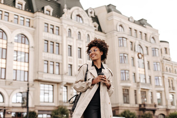 Pretty young happy brunette dark-skinned woman in black dress and trendy beige trench coat smiles and holds coffee cup outdoors.