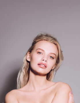 Closeup of pretty young woman face with blue eyes, blonde hair, has nude makeup, perfect soft skin, standing shirtless with bare shoulders, looking at camera. Studio grey background
