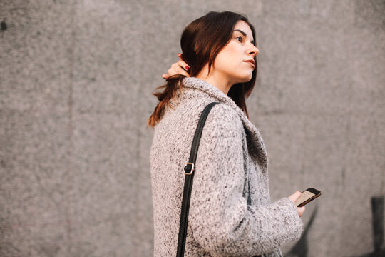 Thoughtful woman holding smart phone walking against gray wall in city