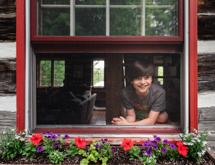 Young happy boy looking out open window of log cabin on summer day.