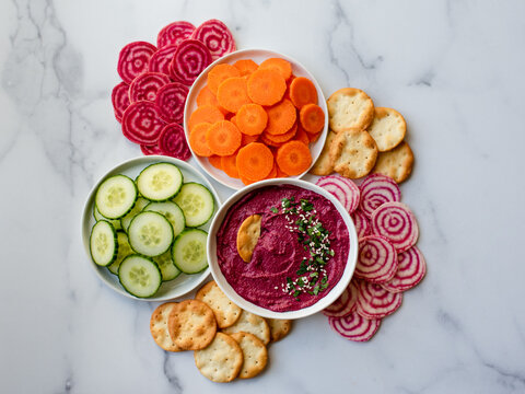 Bowl of beet hummus, vegetables and crackers on white marble counter.