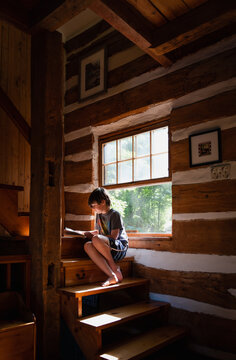 Boy reading a book indoors on the sunny step of a log cabin home.