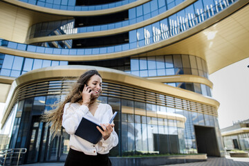 Girl with long hair in city speaks on phone and looks in a notebook.