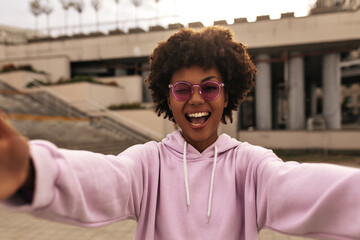 Attractive beautiful curly dark-skinned woman in pink sunglasses and purple hoodie rejoices, winks and takes selfie outside.