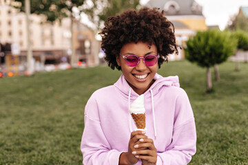 Happy excited African dark-skinned curly woman in fuchsia sunglasses and pink hoodie smiles...