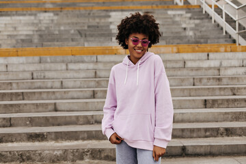 Charming pretty dark-skinned curly brunette woman in pink sunglasses and purple hoodie smiles and...