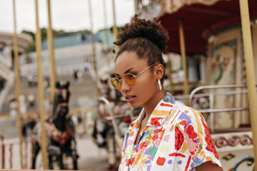 Charming stylish curly dark-skinned woman in colorful fashionable blouse and orange sunglasses poses near carousel.