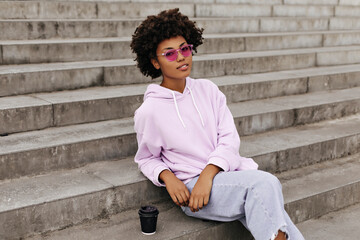 Attractive curly brunette dark-skinned woman in denim pants and purple hoodie looks into camera. African girl in pink sunglasses sits on stairs outdoors.