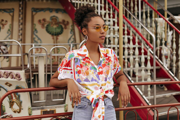 Calm young brunette curly dark-skinned woman in denim pants, colorful blouse and orange sunglasses...