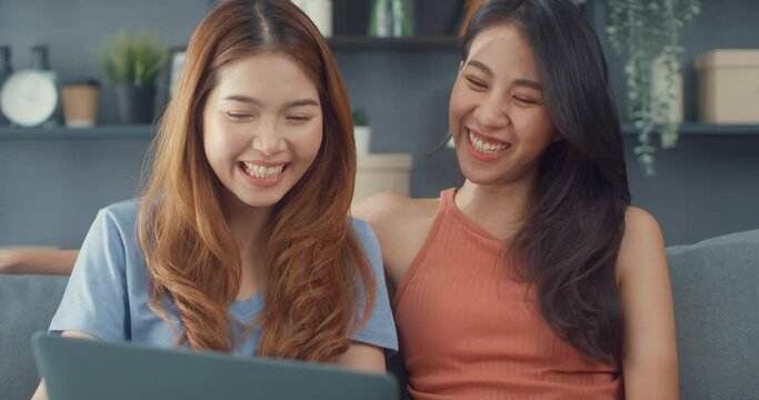 Two Asia lesbian women site on couch together looking at laptop screen in living room at home together. Happy couple roommate ladies enjoy web surfing online shopping, Lifestyle woman at home concept.