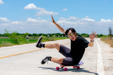 A young Asian man in a black shirt and pants sits on a skateboard. On a country road on a sunny day with sky. Looking at the camera, Play surf skate.