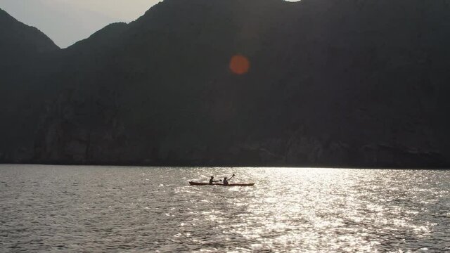Tracking shot of silhouette of two people kayaking in the ocean next to towering limestone fjord mountains. Lens flare at top of frame, sun reflecting on the calm water in Khasab bay Musandam, Oman