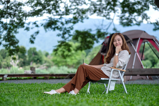 Portrait image of a beautiful young asian woman drinking coffee while sitting on camping chair with a tent in the park