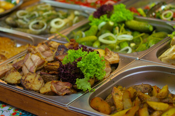 Various products are offered at a street food fair, event, festival. Fried potatoes and other foods on the counter next to large chunks of freshly cooked meat, garnished with bright green lettuce.