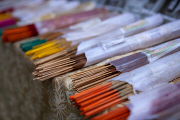 Fototapeta premium Multi-colored orange, yellow, red, green and brown incense sticks lie half visible in white paper wrappers on a counter covered in brown patterned cloth. Blurred background. Close-up.