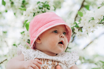 Smiling beautiful small and pretty baby girl. Happy funny and attractive infant girl. Young infant in hat on the blooming apple tree background. Cute baby caucasian ethnicity with blue eyes.