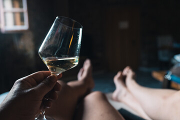 hand with a glass of white wine and legs of a couple lying on a bed in a rural hotel.