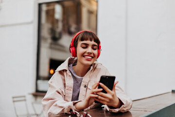 Positive short-haired girl in red headphones smiles outdoors. Brunette woman in beige jacket holding phone and listening to music outside.. - Powered by Adobe