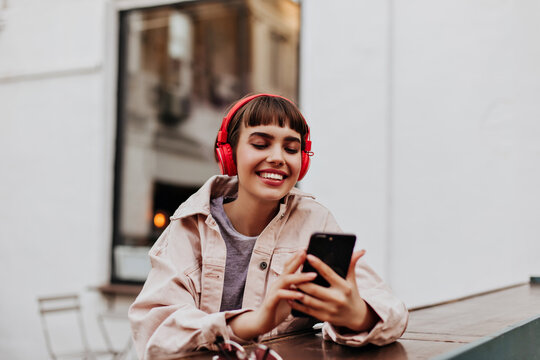 Positive short-haired girl in red headphones smiles outdoors. Brunette woman in beige jacket holding phone and listening to music outside..
